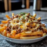 Plate of crispy French fries topped with seasoned ground beef, melted cheddar and mozzarella cheese, and chopped parsley on a granite countertop.